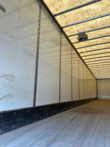 Van trailer interior showing white side walls with vertical supports, wooden floor, and overhead lighting fixture.