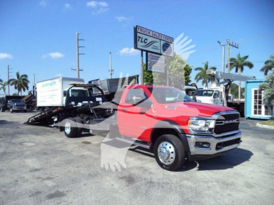 Ram 5500 chassis cab/rollback tow truck in red parked at dealership lot with other trucks and palm trees in background.