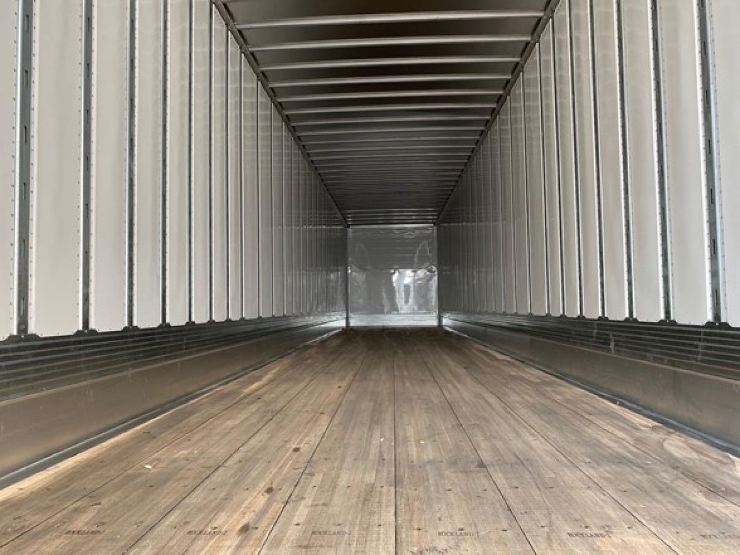 Trailer interior showing empty wooden floor and metal sidewalls and ceiling, long rectangular cargo trailer with ribbed walls.