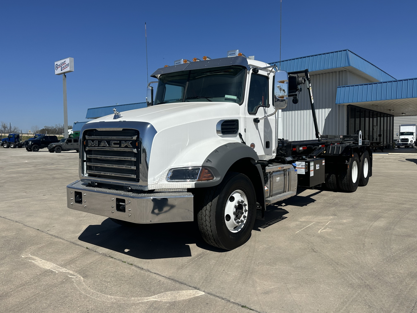 Mack GR64B truck in a dealership lot, front grille with MACK letters, white cab, flatbed roll-off chassis, side mirrors, and chrome bumper.