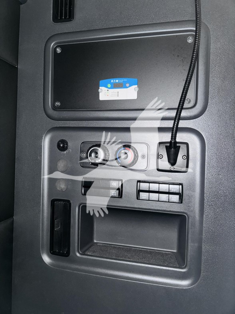 Dash panel of Freightliner Cascadia showing control knobs, a USB/communication port, and a storage pocket; top center shows a small blue-and-white label in a recessed compartment.