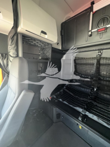Truck cab interior of a 2023 Freightliner Cascadia 126 sleeper showing bunk area with storage nets and partition, driver seat visible.