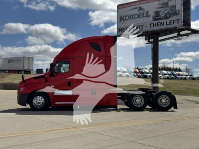 Truck cab and trailer chassis of a Freightliner Cascadia 126 in red with white graphic, parked in lot; billboard in background.