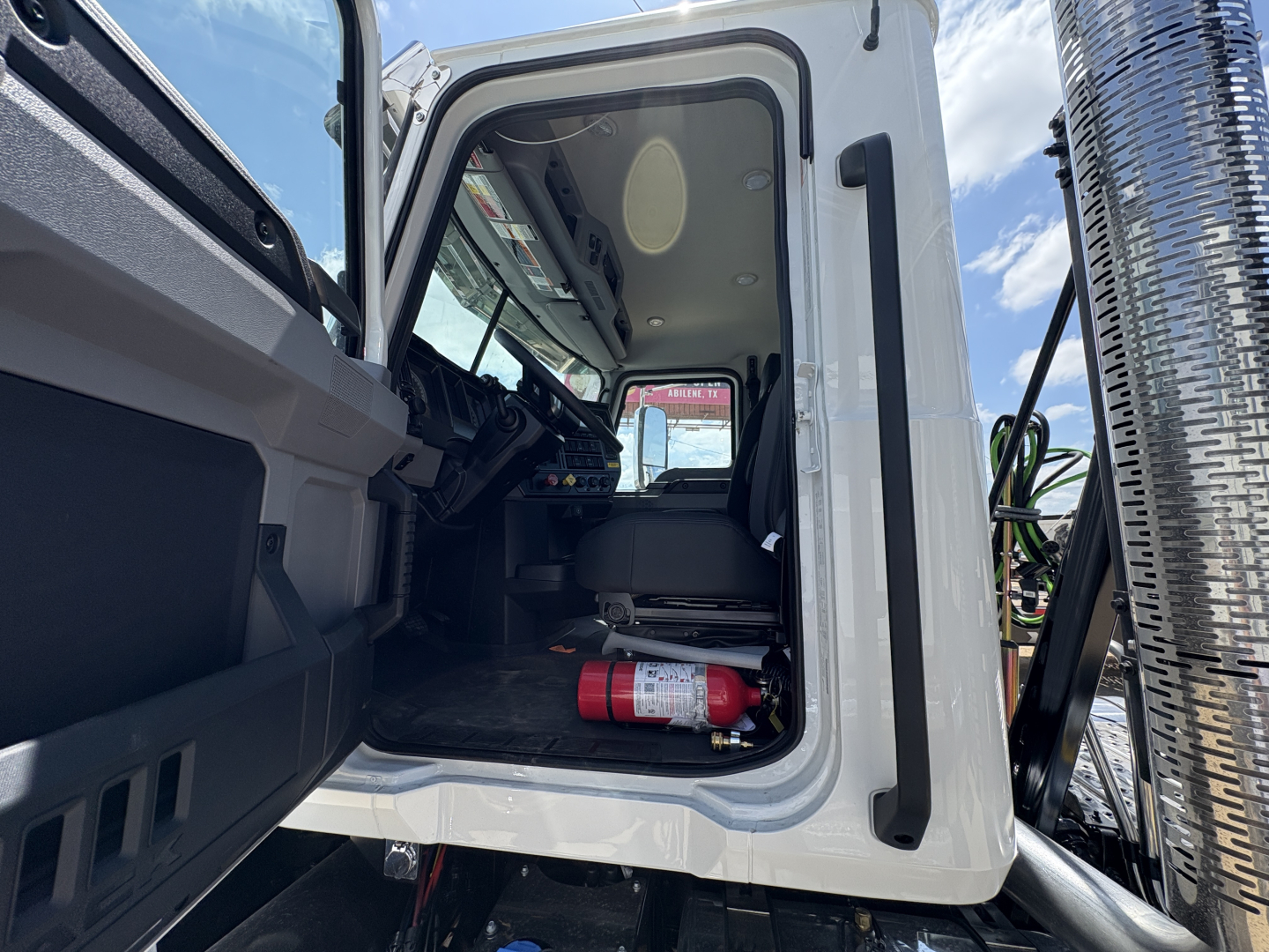 Cab interior of a new 2027 Mack PI64T Daycab truck with open driver's door showing seating, dashboard, and a red fire extinguisher on the floor.