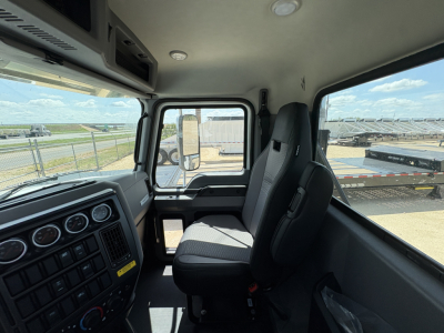 Cab interior of a new 2027 Mack PI64T daycab showing steering wheel dashboard, gauges, and front seats with view through windshield to yard.