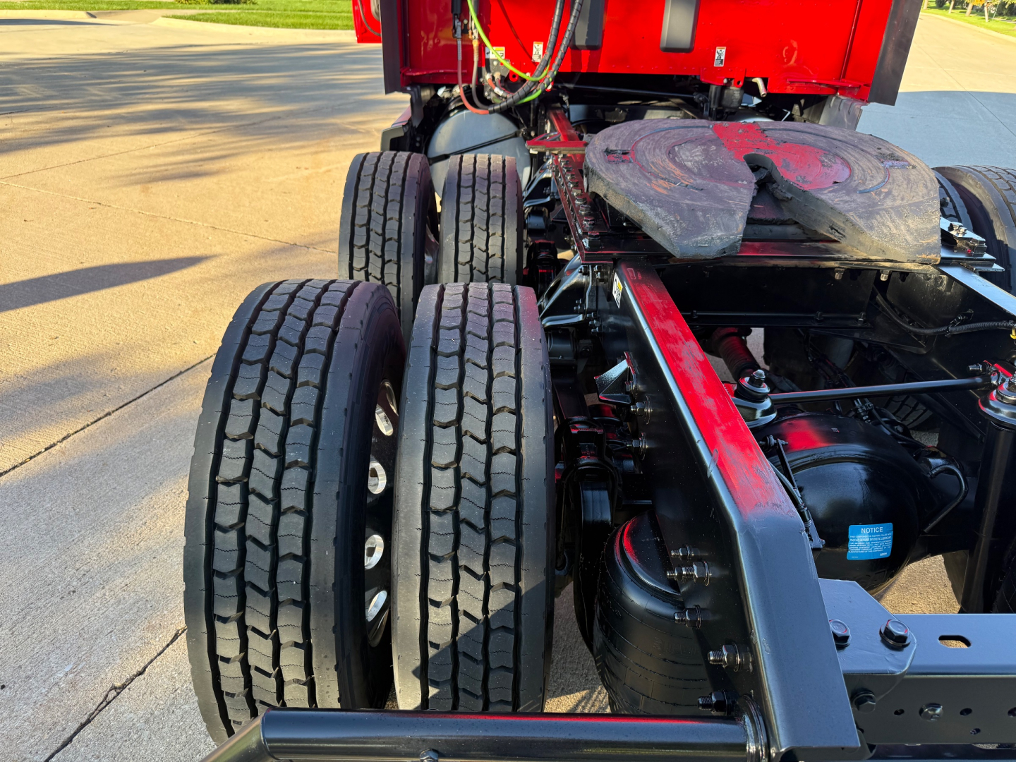 Kenworth T680 rear chassis view showing dual rear axles and tires with drivetrain components visible, red sleeper cab in background.