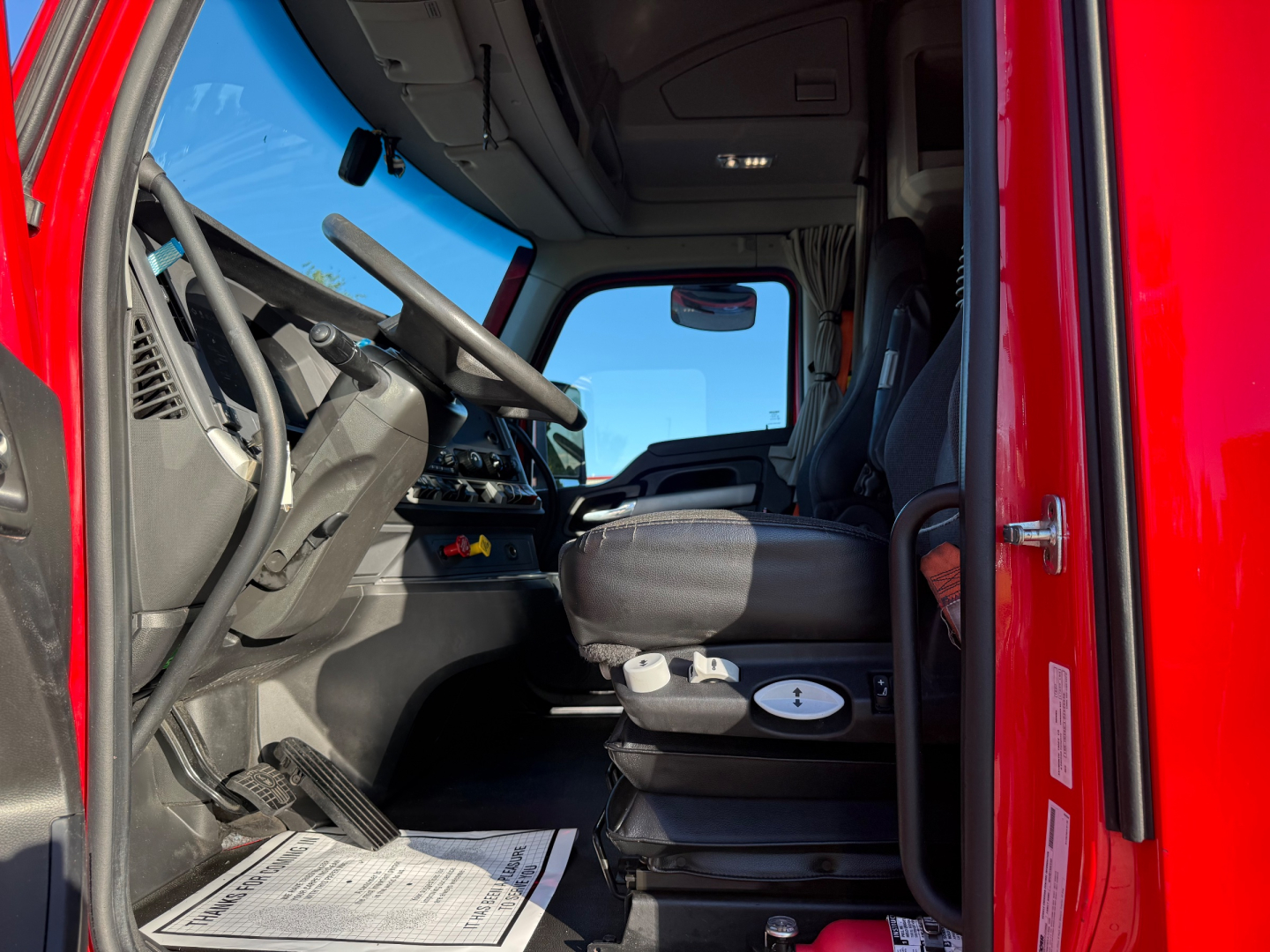 Truck cab interior view of a red 2023 Kenworth T680, with dashboard, steering wheel, driver seat, and center console visible.