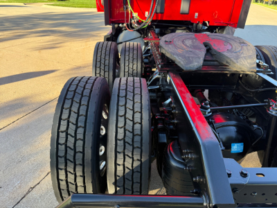 Kenworth T680 rear chassis view showing dual rear axles and tires with drivetrain components visible, red sleeper cab in background.