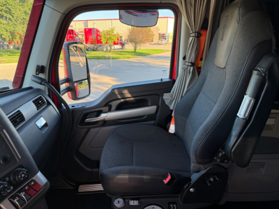 Cab interior of a 2023 Kenworth T680 daytime view showing driver seat and part of dashboard. Side mirror visible through window.