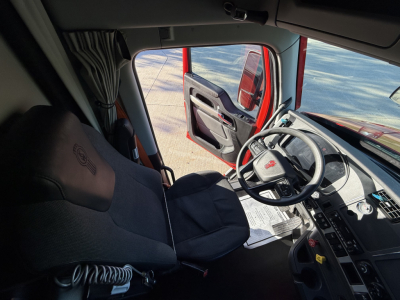 Truck cab interior of Kenworth T680, driver seat, steering wheel, dashboard visible with door open, red exterior visible through window.