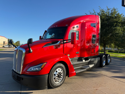 Red Kenworth T680 semi-truck in a parking lot, front-left view, with grille and sleeper cab visible.