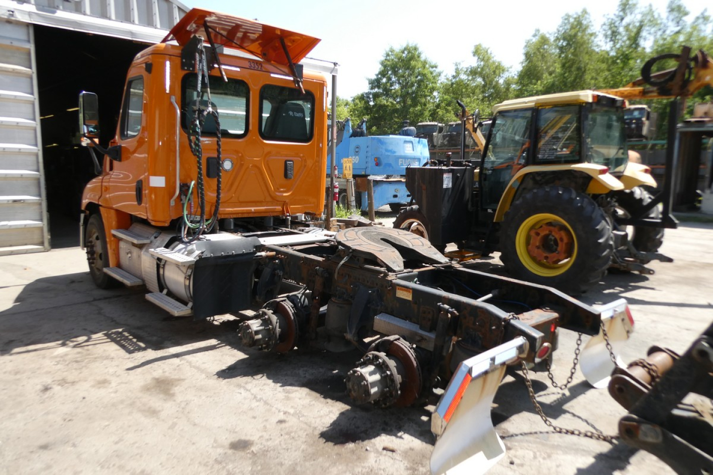 Orange Freightliner Cascadia 125 cab with exposed chassis and front axle components, yard photo with other construction equipment in background.