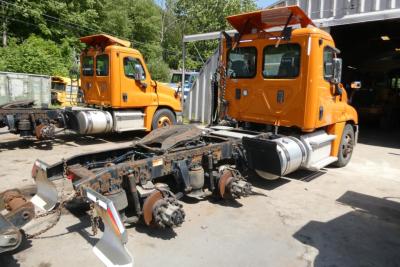 Two orange Freightliner Cascadia 125 trucks without cabs in a yard; detached suspension and drivetrain components on the ground in front; side view shows cab doors and windows.
