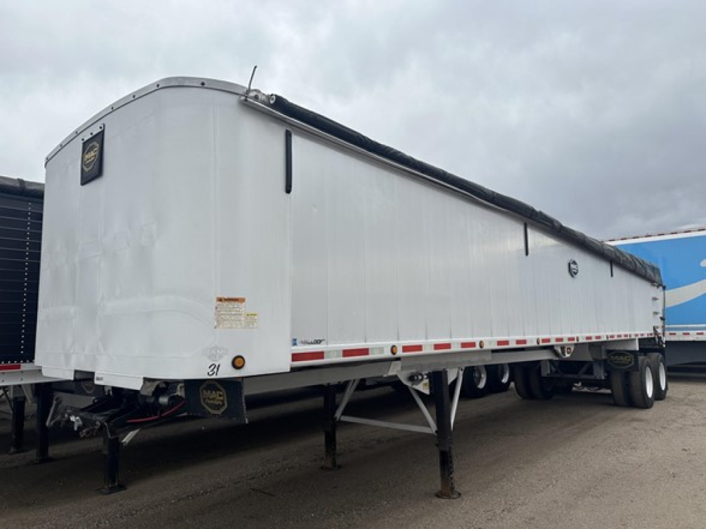 Framed aluminum end dump trailer showing elevated front with pin hitch, landing gear, and dual rear axles; white exterior with black tarp rail and warning labels.