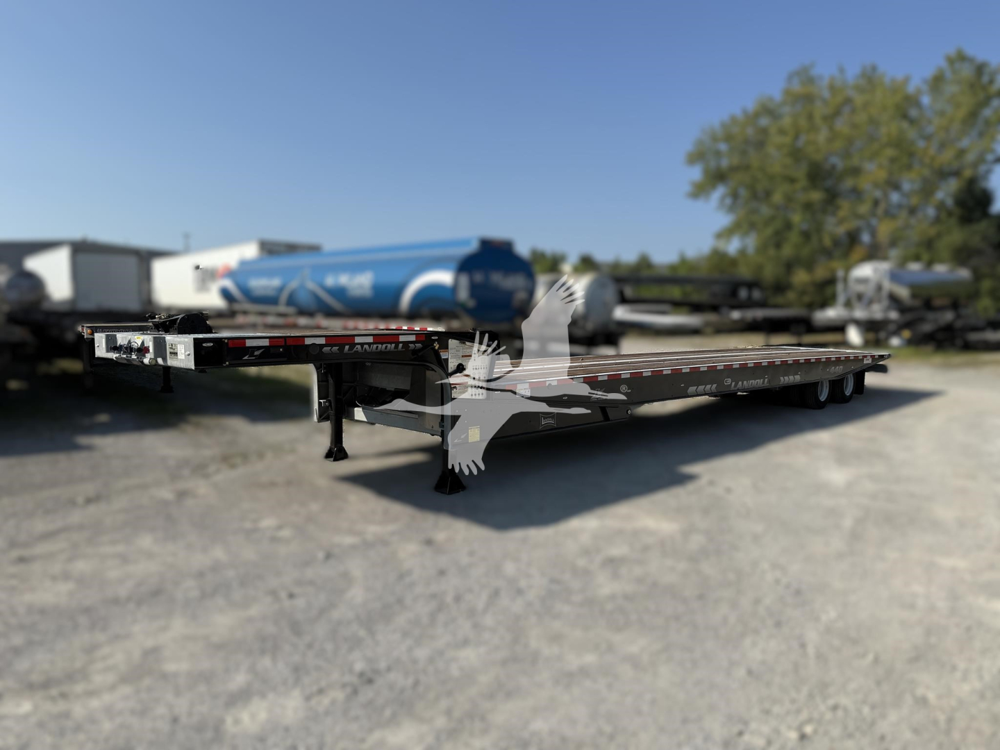 Landoll 440 traveling axle trailer showing front left corner with metal deck and support legs. Text on side reads ‘LANDOLL’ and other small labels; background includes parked trailers and a blue tanker.