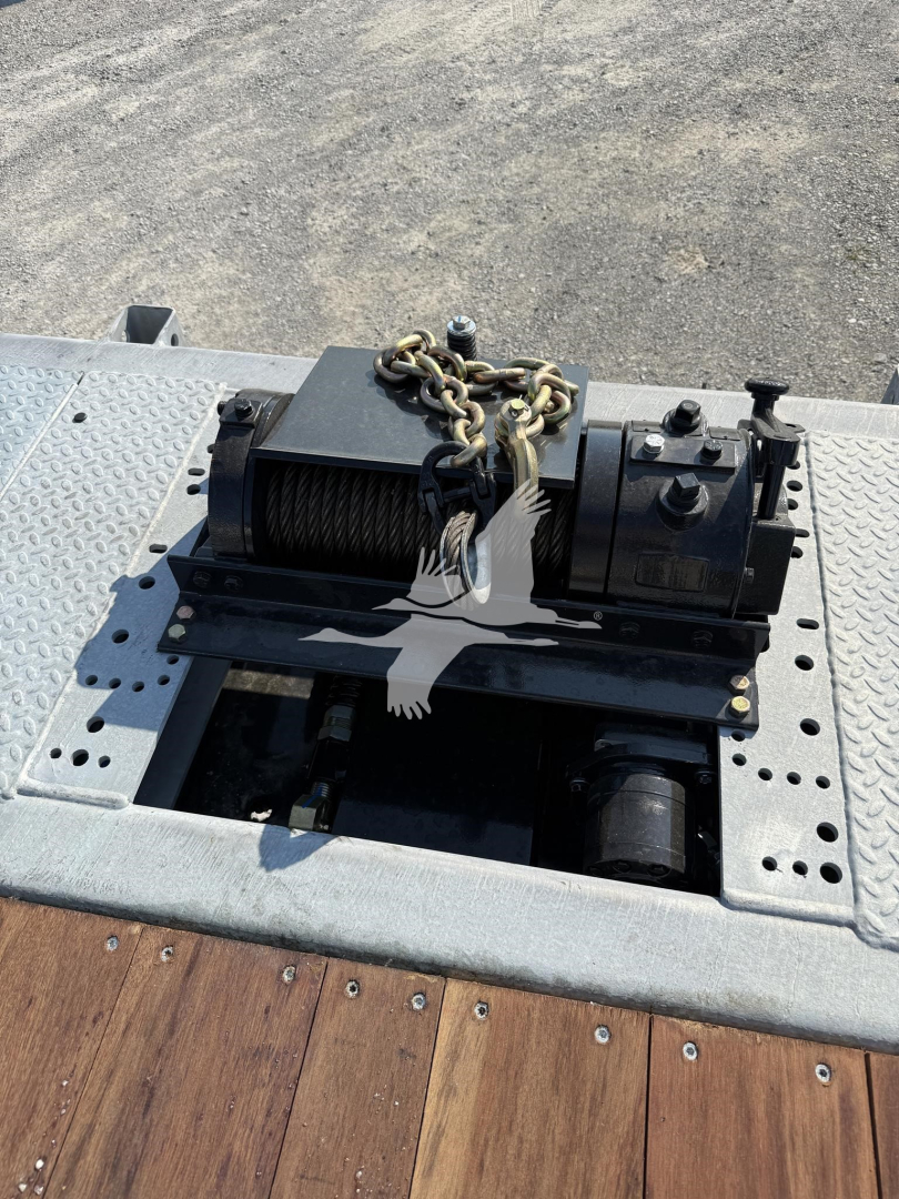 Winch assembly mounted on trailer deck with chain, motor, and spool; visible bolts and perforated steel plate; no readable serials or model numbers visible on this shot.
