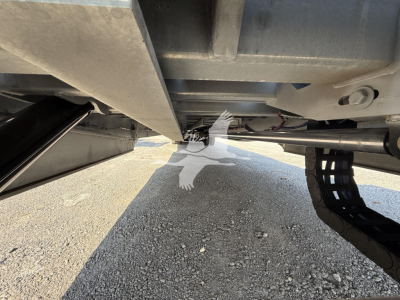 Underbelly of a trailer showing frame and landing gear with a chain and hose; ground visible beneath. No legible label text visible in this shot.