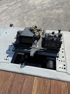 Winch assembly mounted on trailer deck with chain, motor, and spool; visible bolts and perforated steel plate; no readable serials or model numbers visible on this shot.