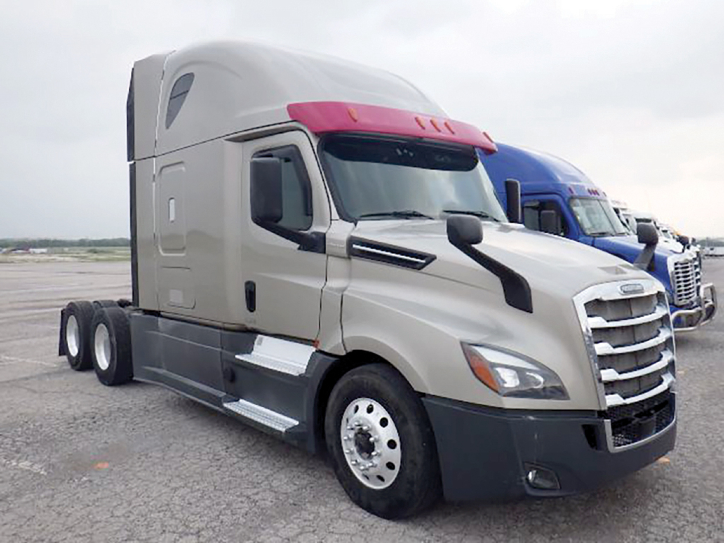 Freightliner Cascadia126 sleeper cab truck in a parking lot, front and side view, chrome grille, red sun visor, light gray cab.
