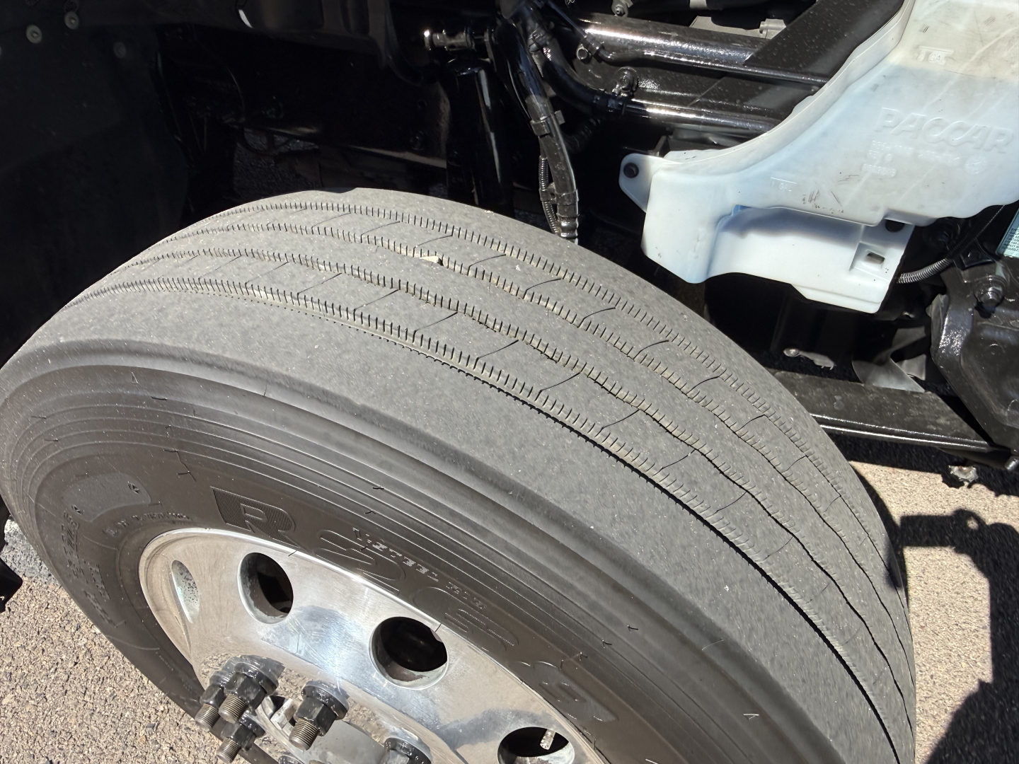 Front left wheel of a 2023 Kenworth T680 conventional sleeper truck showing tire and wheel; partial chassis and coolant reservoir visible.