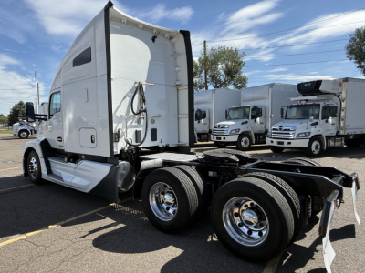 Kenworth T680 tractor in a yard, side view showing rear dual axles, fifth wheel, and white cab with sleeper, chrome wheels, and exposed frame.