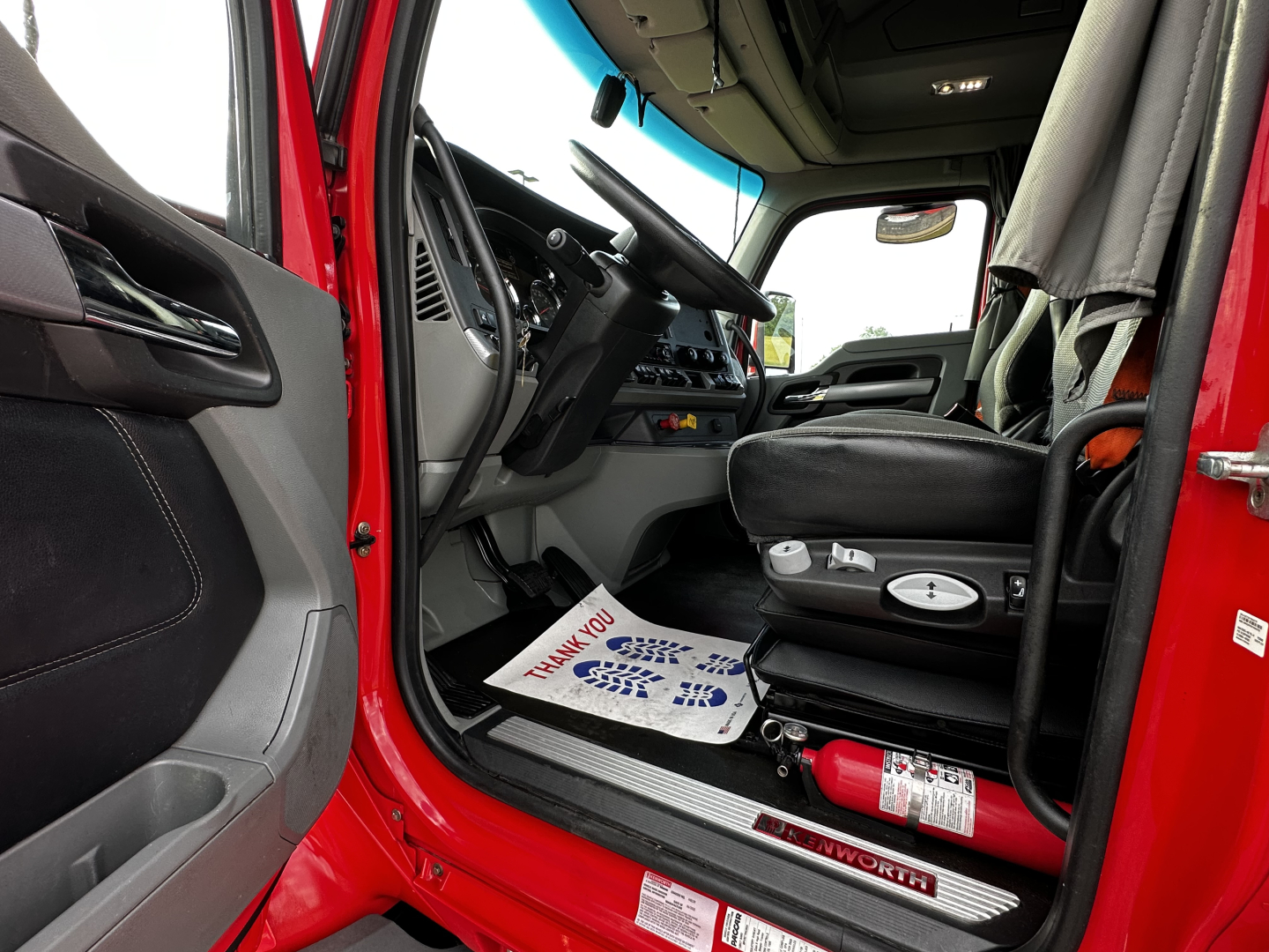 Truck cab interior of a 2022 Kenworth T680 showing the driver's seat, steering wheel, and entry view with a floor mat and a red exterior.