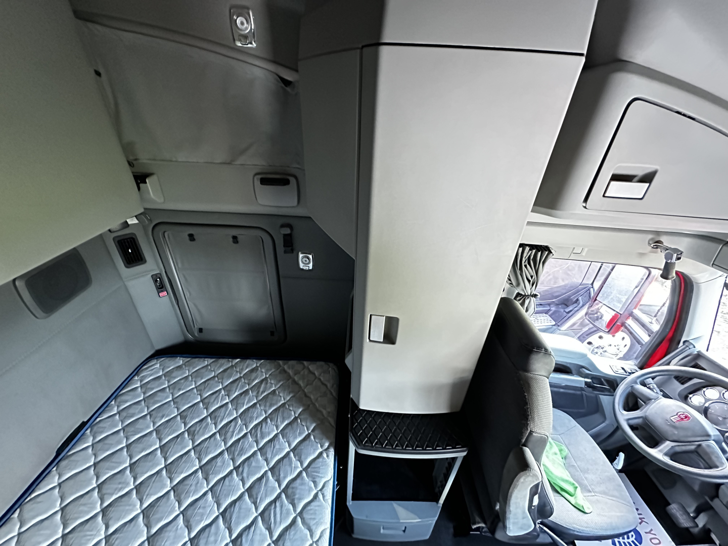 Cab interior of a 2022 Kenworth T680 sleeper showing a vertical center console, driver's seat, dashboard, and a quilted sleeper berth area.
