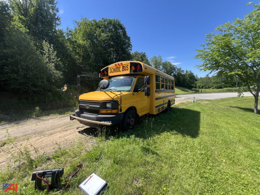 Yellow school bus parked on grass with front view, Chevrolet Express style, 'SCHOOL BUS' sign on top, trees and blue sky in background.