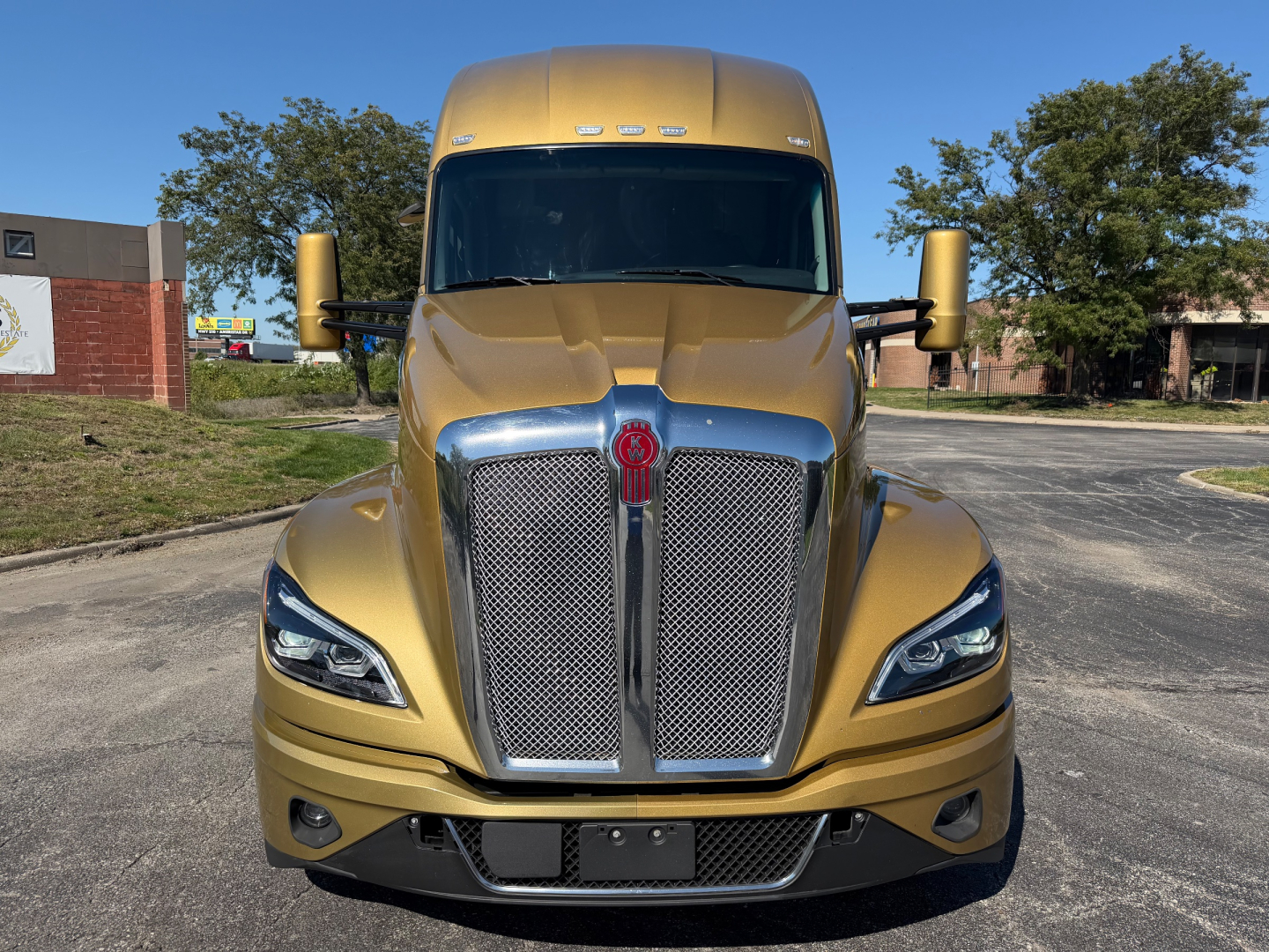 Kenworth T680 front view showing gold exterior, large chrome grille, black bumper, and LED headlights.
