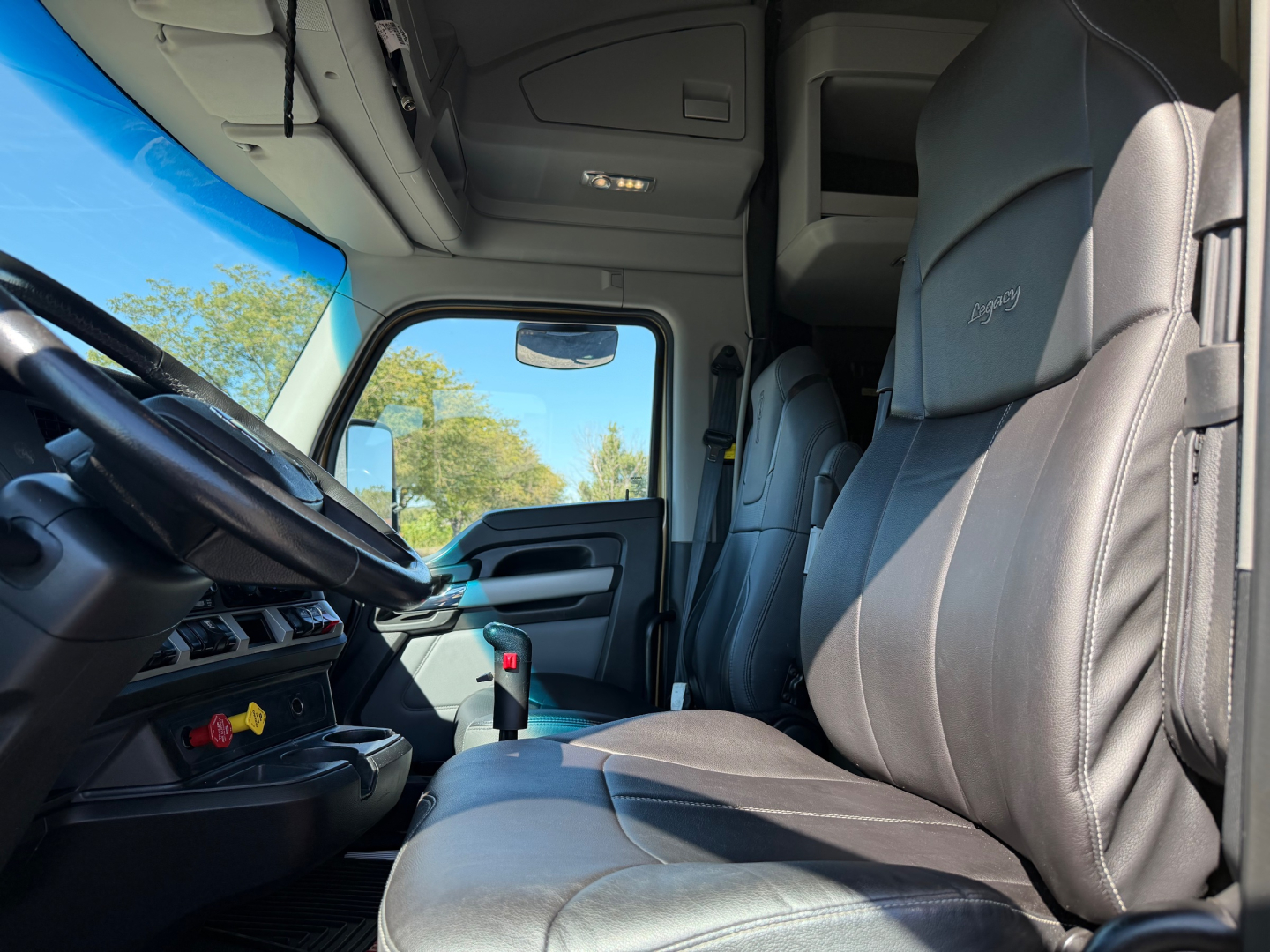 Interior cab of a 2023 Kenworth T680, driver seat and dashboard visible, leather seats, steering wheel, window, overhead storage, modern controls.