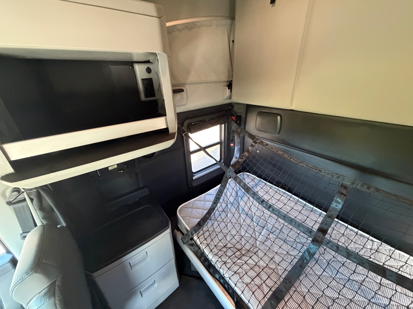 Inside a Kenworth T680 sleeper cab showing a netted bunk area, overhead cabinet, and window; compact dashboard area visible.
