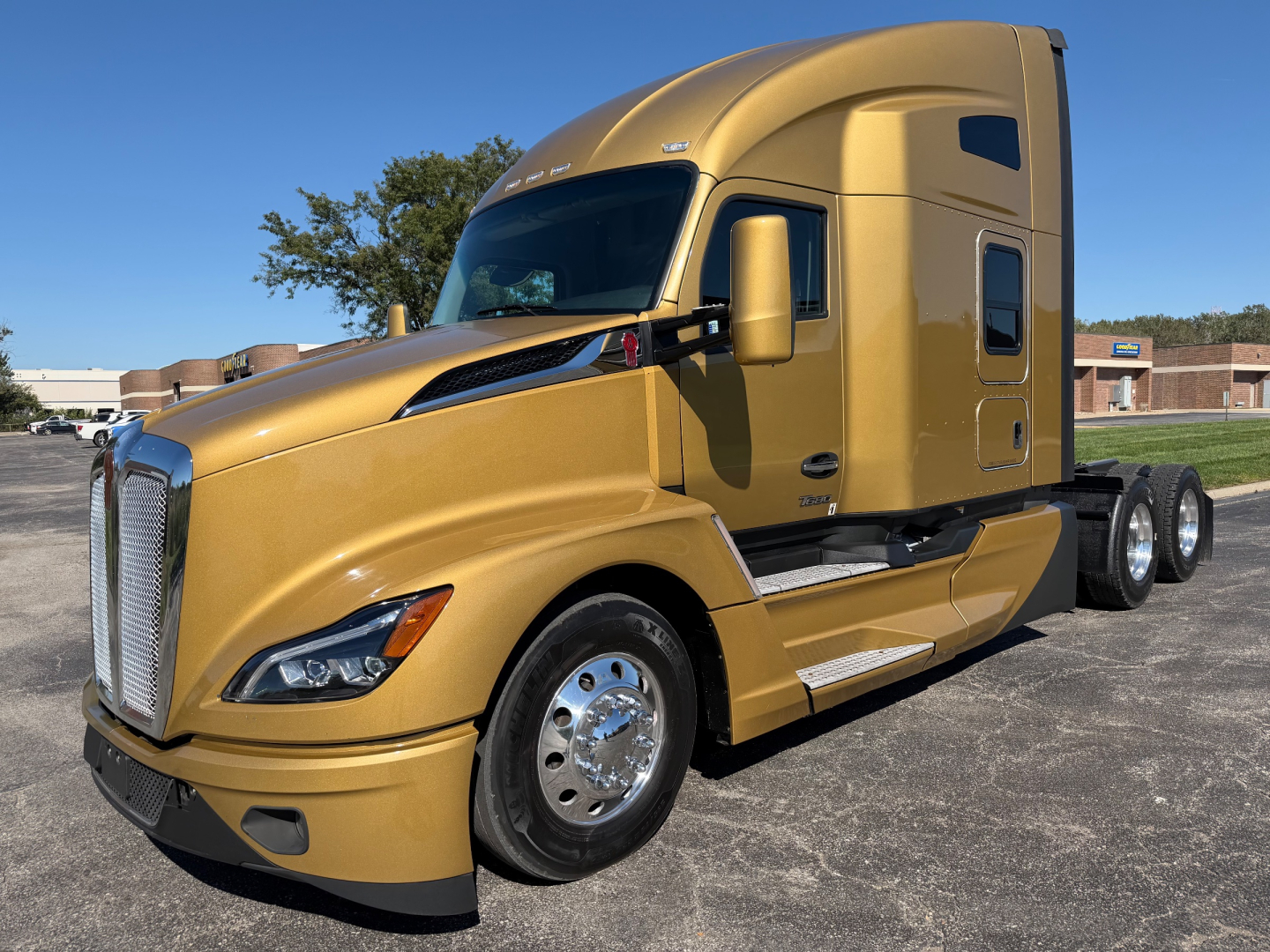 Kenworth T680 tractor in gold color, front left view showing cab and sleeper, with chrome wheels and side steps.