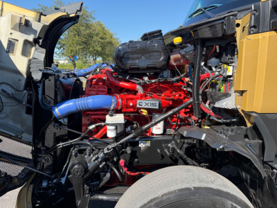 Kenworth T680 engine bay with red engine, blue coolant hose, black battery tray, and various hoses; visible engine components; no plate text legible.