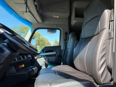Interior cab of a 2023 Kenworth T680, driver seat and dashboard visible, leather seats, steering wheel, window, overhead storage, modern controls.