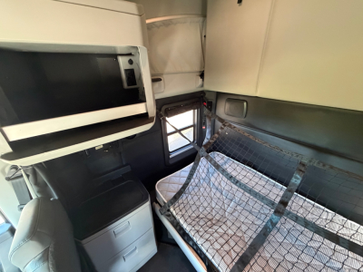 Inside a Kenworth T680 sleeper cab showing a netted bunk area, overhead cabinet, and window; compact dashboard area visible.