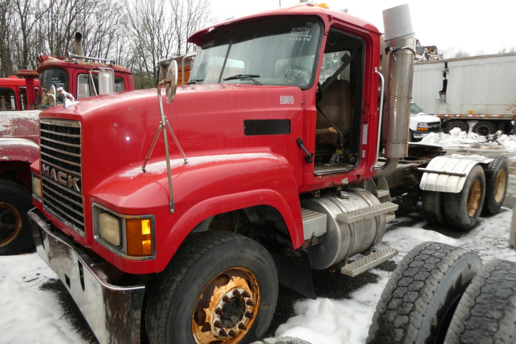 Mack CHN613 conventional daycab truck in red showing front grille and cab, with snow on ground and other trucks in background.