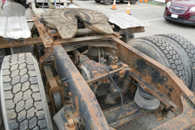 Truck chassis rear view showing rusty frame and drivetrain undercarriage with fifth wheel and tires.