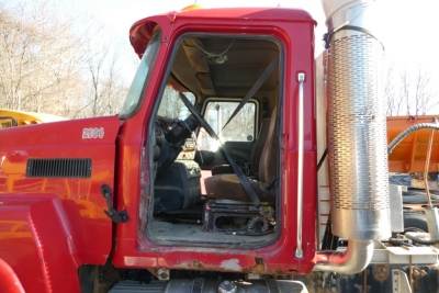 Mack CHN613 cab interior view with red exterior. Door open showing driver seat, steering wheel, and dashboard. Large vertical exhaust stack on the right side. Sticker text not fully legible.