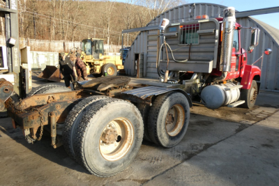 Mack CHN613 conventional daycab truck chassis showing dual rear axles and exposed drive components with a red sleeper cab in background.