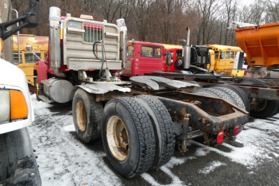 Mack CHN613 truck chassis in yard showing fifth wheel and dual rear axles with tires, container and other trucks in background.