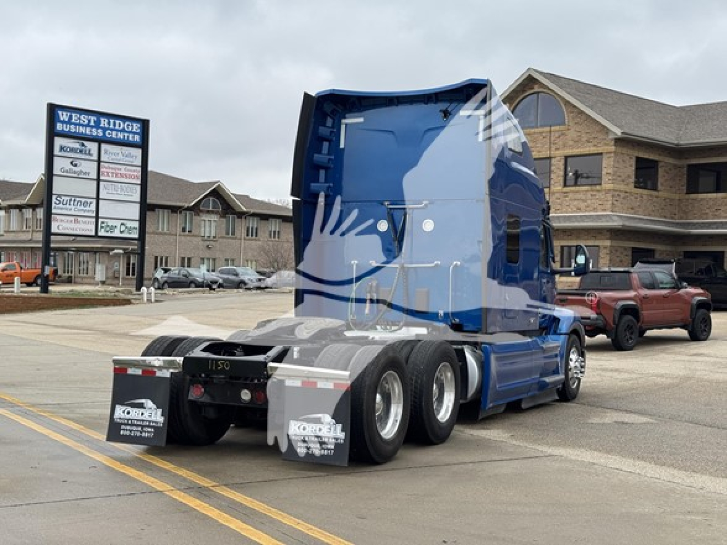 Truck rear view of a blue 2023 Peterbilt 579 tractor in a dealership lot showing third axle and mud flaps with KORBEL logos.