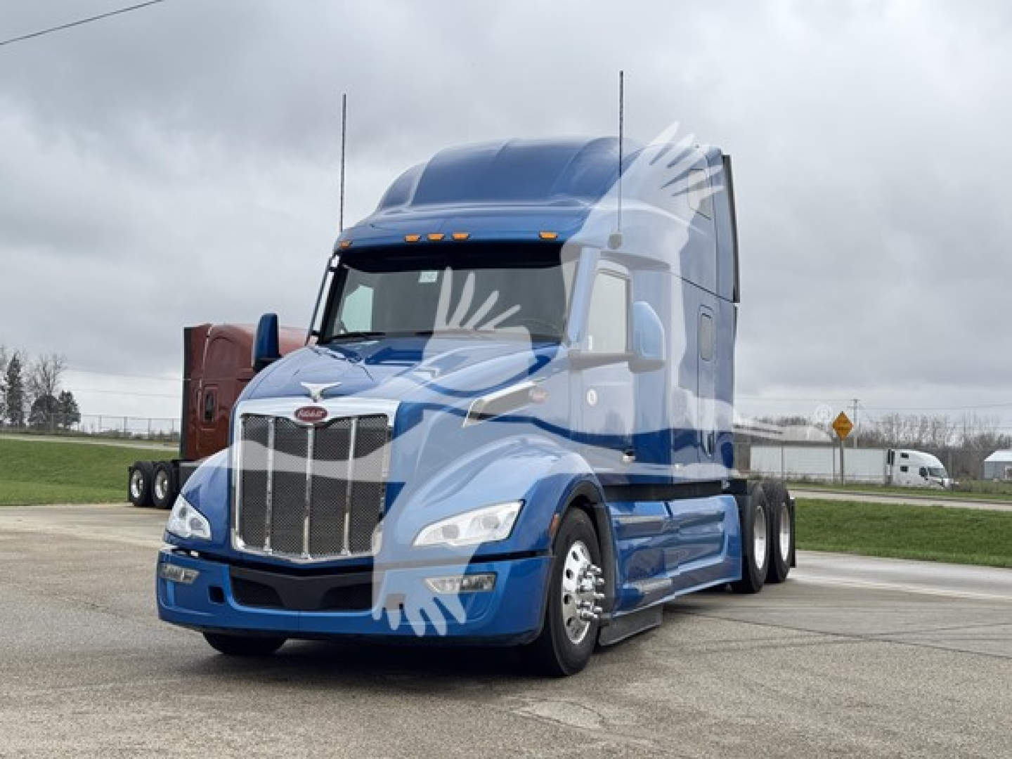 Peterbilt 579 truck in blue exterior, front view, large grille, sleeper cab, two tall antennae visible, cloudy sky.