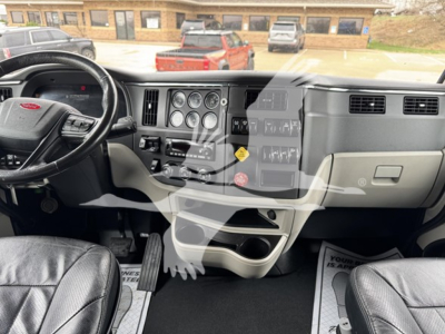 Truck cab interior dash with steering wheel and gauges; center console with switches; Peterbilt emblem on steering wheel; view into sleeper area and front windshield.