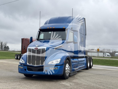 Peterbilt 579 truck in blue exterior, front view, large grille, sleeper cab, two tall antennae visible, cloudy sky.