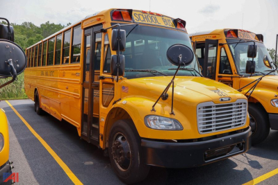 Front and side view of a yellow Freightliner Thomas Saf-T-Liner school bus with 'SCHOOL BUS' signage on top, adjacent bus in background.