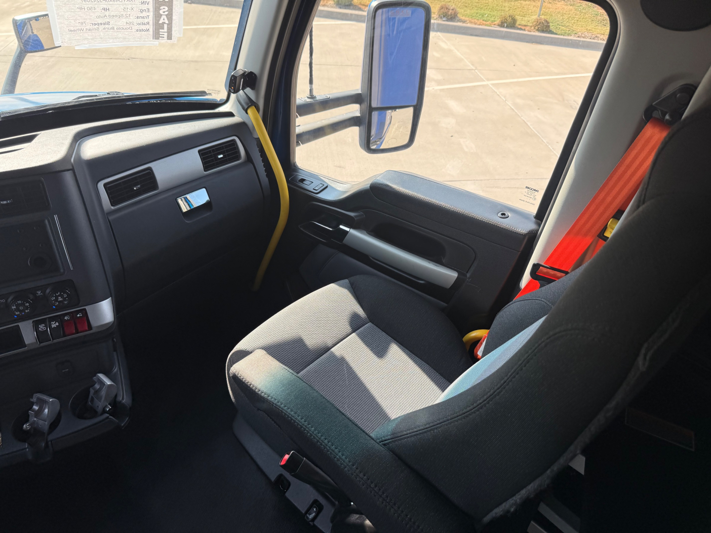 Truck cab interior of a Kenworth T680 showing steering wheel, dashboard, and front passenger seat with orange seat belt in view.