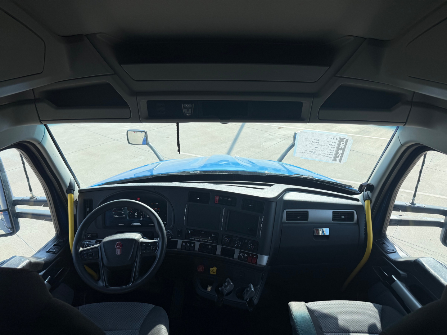 Kenworth T680 cab interior from driver perspective showing steering wheel, dashboard controls, and windshield; door frames with yellow grab handles.