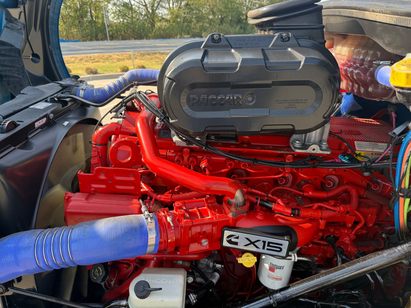 Kenworth T680 engine bay with red engine block, blue intercooler hoses, and D13? The cover shows PACCAR with ZX15 badge on a component; visible components include hoses, electrical wiring, and coolant reservoir.