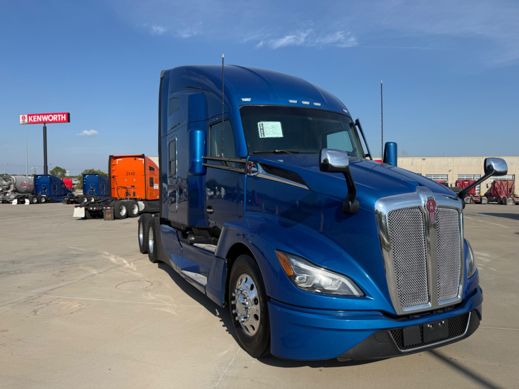 Kenworth T680 truck in blue, front and driver side view, large grille, badge, chrome mirrors, daytime sky.