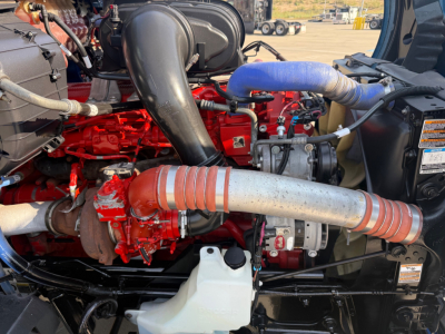 Kenworth T680 engine bay with red engine, large black intake, silver exhaust, blue hose, white coolant reservoir, and various hoses and components visible.